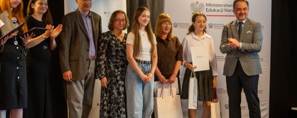 Grupa osób stojąca na scenie przed banerami z napisami „Archiwa Państwowe” i „Ministerstwo Edukacji Narodowej”, w środku dwie młode dziewczyny trzymające torby upominkowe i dokument, dorośli po bokach – po prawej mężczyzna w garniturze klaszcze, po lewej kobieta trzyma mikrofon i notatki.