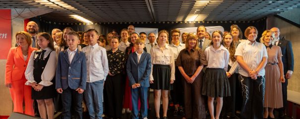 Grupa dzieci, młodzieży i dorosłych ustawiona w kilku rzędach na scenie przed czarną kurtyną, wszyscy zwróceni ku aparatowi; u góry widoczna metalowa kratownica sufitu z liniami świetlnymi, na pierwszym planie formalne stroje (marynarki, białe koszule, sukienki) i po lewej duży głośnik.