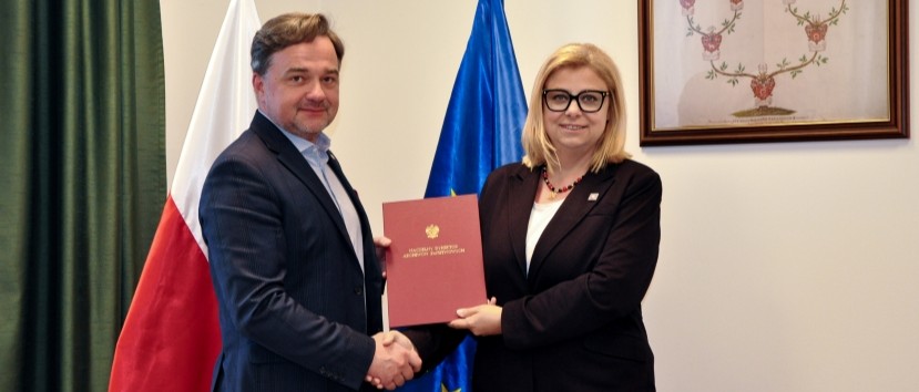 Dwie osoby — mężczyzna w ciemnym garniturze i kobieta w czarnym żakiecie i okularach — stoją w pomieszczeniu przed flagami Polski i Unii Europejskiej, podają sobie ręce i wspólnie trzymają czerwony dokument z widocznym złotym godłem, a na ścianie obok wisi oprawiony obraz.