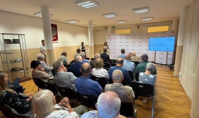 Sala z rzędami siedzącej publiczności zwróconej ku mównicy, gdzie dwie kobiety przemawiają obok ekranu z napisem Fotografie z Archiwum ANDRZEJA FRISZKIEGO, z gablotą ekspozycyjną po lewej, polskim herbem i krzyżem na ścianie, kilkoma filarami i sufitowymi panelami świetlnymi nad drewnianą podłogą.