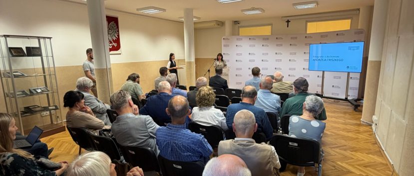 Sala z rzędami siedzącej publiczności zwróconej ku mównicy, gdzie dwie kobiety przemawiają obok ekranu z napisem Fotografie z Archiwum ANDRZEJA FRISZKIEGO, z gablotą ekspozycyjną po lewej, polskim herbem i krzyżem na ścianie, kilkoma filarami i sufitowymi panelami świetlnymi nad drewnianą podłogą.