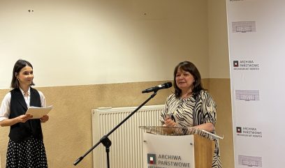 Dwie kobiety na sali: po prawej przy mównicy kobieta w bluzce w czarno-białe paski stojąca przy mikrofonie, po lewej kobieta trzymająca dokumenty w białej koszuli, czarnym kamizelce i spódnicy w groszki; w tle beżowa ściana, grzejnik i stojący roll-up z logo 