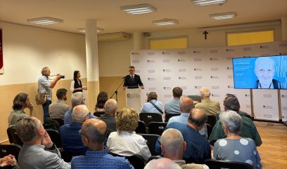 Sala konferencyjna z grupą uczestników siedzących tyłem do kamery, mężczyzną przemawiającym przy mównicy przed ścianką z logo „Archiwa Państwowe”, obok fotografem z aparatem i kobietą z notatkami, po prawej dużym ekranem wyświetlającym portret i informacje oraz krzyżem na ścianie i widocznymi sufitowymi oprawami świetlnymi.