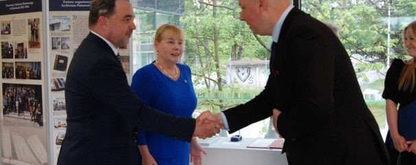 Trzy osoby w jasnym pomieszczeniu przy dużych oknach: po lewej mężczyzna w ciemnym garniturze ściska dłoń mężczyzny w garniturze po prawej, między nimi stoi kobieta w niebieskiej sukience, w tle plansze wystawy z fotografiami i widoczne przez okno drzewa oraz zabudowania.