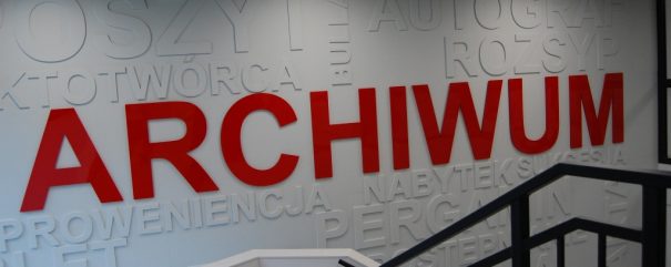 Wnętrze z białą ścianą pokrytą dużymi czerwonymi literami ARCHIWUM i wypukłymi białymi napisami w tle, czarną metalową balustradą schodów na pierwszym planie oraz sufitem z listwami i lampami.