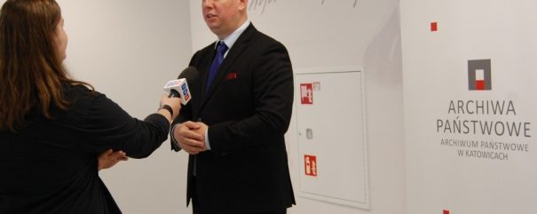 Mężczyzna w ciemnym garniturze i niebieskim krawacie stoi przy ścianie, naprzeciw niego kobieta z długimi brązowymi włosami trzyma ku niemu mikrofon, po prawej widoczny roll‑up z napisem 