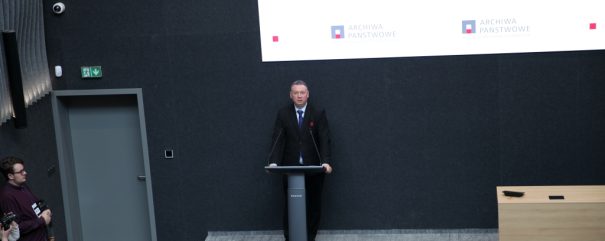 Mężczyzna w ciemnym garniturze stoi za mównicą z dwoma mikrofonami na podwyższeniu przed ciemnoszarym panelem z białym banerem z logo 