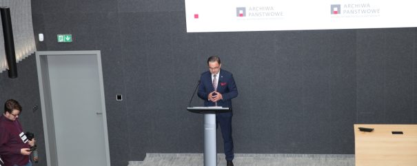 Mężczyzna w garniturze stoi za mównicą na podwyższeniu przed dużą szarą ścianą z powtarzającym się logo 