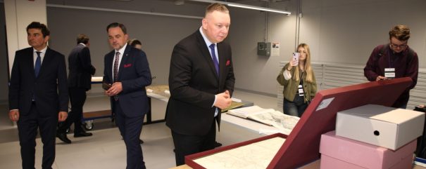 Kilka osób w jasnym pomieszczeniu przy stole z rozłożonymi mapami i pudełkami — mężczyzna w ciemnym garniturze pochylony nad oprawioną mapą na czerwonym podwyższeniu, obok inni mężczyźni w garniturach, kobieta robiąca zdjęcie telefonem i mężczyzna z identyfikatorem 