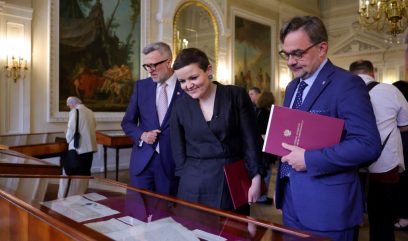 Troje dorosłych osób — kobieta w czarnej sukience i dwaj mężczyźni w niebieskich garniturach — stoi przy drewniano-szklanej gablocie z dokumentami w ozdobnej, dobrze oświetlonej sali z dużymi obrazami na ścianach i kryształowym żyrandolem, każdy z nich trzyma czerwoną teczkę.