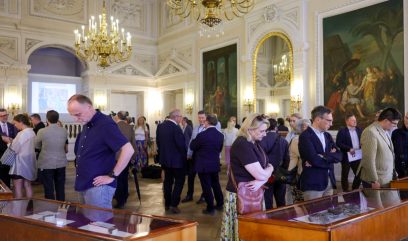 Wnętrze bogato zdobionej, klasycystycznej sali z ozdobnym sufitem, lustrami i dwoma dużymi obrazami na ścianach, podwisającymi kryształowymi żyrandolami, na środku której grupy ludzi w formalnym i casualowym ubraniu oglądają przedmioty wystawione w przeszklonych gablotach.