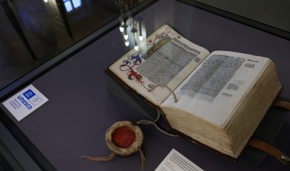 Otwarte, grube, iluminowane księgi leżące na fioletowym podłożu w szklanej gablocie, z ozdobną inicjałą i gęstym czarnym tekstem, obok okrągłego pojemnika z czerwoną woskową pieczęcią i sznurkami oraz kartą z logo UNESCO i odbiciami lamp na szybie.