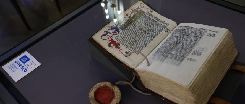 Otwarte, grube, iluminowane księgi leżące na fioletowym podłożu w szklanej gablocie, z ozdobną inicjałą i gęstym czarnym tekstem, obok okrągłego pojemnika z czerwoną woskową pieczęcią i sznurkami oraz kartą z logo UNESCO i odbiciami lamp na szybie.