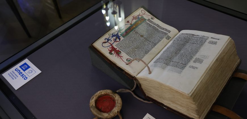 Otwarte, grube, iluminowane księgi leżące na fioletowym podłożu w szklanej gablocie, z ozdobną inicjałą i gęstym czarnym tekstem, obok okrągłego pojemnika z czerwoną woskową pieczęcią i sznurkami oraz kartą z logo UNESCO i odbiciami lamp na szybie.
