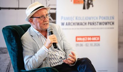 Mężczyzna w kapeluszu z czarną wstążką i okularach, ubrany w jasną marynarkę i koszulę w pionowe paski, siedzi na zielonym aksamitnym fotelu, trzyma mikrofon i gestykuluje dłonią, na rozmytym tle widoczny plakat z polskim tekstem.
