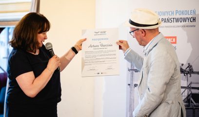 Kobieta z mikrofonem po lewej i mężczyzna w jasnym garniturze i słomkowym kapeluszu po prawej trzymają razem certyfikat z widocznym nagłówkiem PODZIĘKOWANIA na jasnym tle