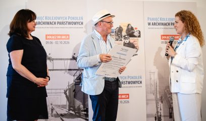 Trzy osoby stoją na tle banerów z grafikami kolejowymi i napisem „Pamięć Kolejowych Pokoleń w Archiwach Państwowych” — mężczyzna w środku w jasnej marynarce, okularach i kapeluszu trzyma dokument z nagłówkiem „Podziękowania”, kobieta po prawej w białym garniturze trzyma mikrofon i mówi, a kobieta po lewej w ciemnej sukience stoi złożonymi rękami.
