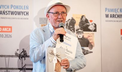 Mężczyzna w białym kapeluszu i okularach, ubrany w jasną paskowaną marynarkę, trzyma mikrofon, dokument i ozdobne pudełko z kokardą, przemawiając przed planszą z polskim tekstem i starymi zdjęciami.