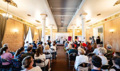 Duża ozdobna sala z tapetami, zasłonami i żyrandolami, z wysokimi białymi kolumnami i sufitem z projektorem; publiczność siedzi w rzędach składanych krzeseł zwrócona ku małej scenie z dwiema osobami siedzącymi przy stoliku, plakatami informacyjnymi i kamerą na statywie po prawej.