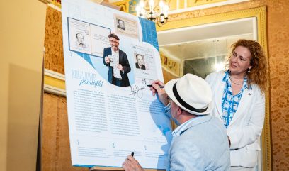 Mężczyzna w jasnej marynarce i słomkowym kapeluszu podpisuje dużą planszę na sztaludze z grafikami i zdjęciami dokumentów zatytułowaną „Kolejowe pamiątki”, obok stoi kobieta w białej marynarce i niebieskiej bluzce, a w tle widać ozdobną tapetę, duże lustro i wiszący żyrandol.