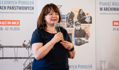 Kobieta w ciemnym topie i krótkich brązowych włosach trzyma mikrofon, z widocznym naszyjnikiem, bransoletką i zegarkiem, stojąca na tle banerów z polskim tekstem oraz kolażem historycznych zdjęć i grafik pociągów.