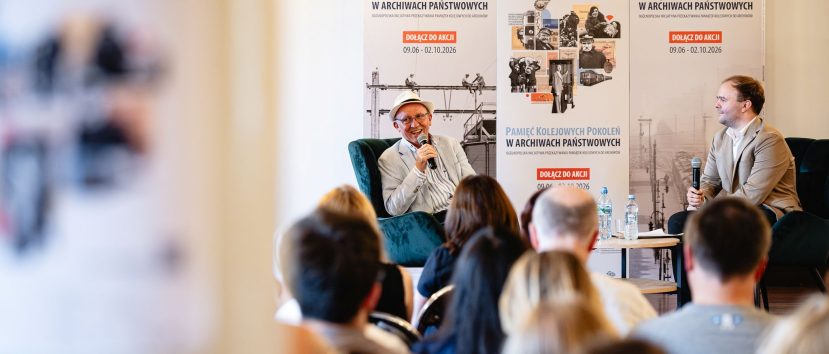 Dwóch mężczyzn siedzi na krzesłach podczas rozmowy na scenie — mężczyzna po lewej w jasnym garniturze i kapeluszu trzyma mikrofon, mężczyzna po prawej w beżowej marynarce też trzyma mikrofon, za nimi duże plakaty z napisem Pamięć Kolejowych Pokoleń w Archiwach Państwowych i informacją o datach, przed nimi rozmyta publiczność oraz mały stolik z butelkami wody.