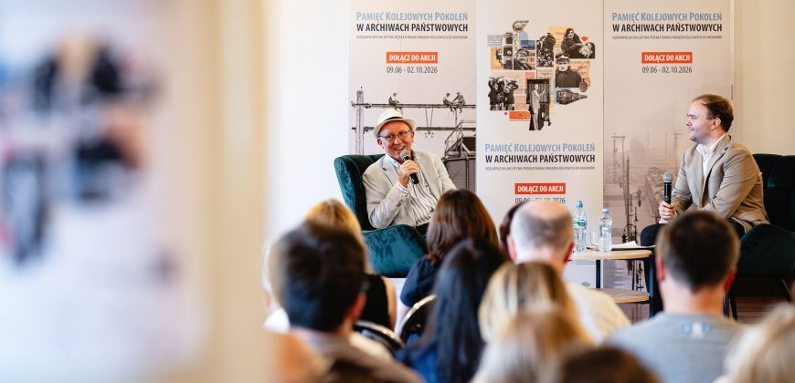 Dwóch mężczyzn siedzi na krzesłach podczas rozmowy na scenie — mężczyzna po lewej w jasnym garniturze i kapeluszu trzyma mikrofon, mężczyzna po prawej w beżowej marynarce też trzyma mikrofon, za nimi duże plakaty z napisem Pamięć Kolejowych Pokoleń w Archiwach Państwowych i informacją o datach, przed nimi rozmyta publiczność oraz mały stolik z butelkami wody.