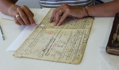 Dłonie osoby w paskowanej koszulce na białym stole dotykające bawełnianym patyczkiem pożółkłe, zapisane drobnym pismem karty z czerwonym okrągłym stemplem; widoczne pierścionki na palcach i cienka bransoletka, po lewej stalowe narzędzie, po prawej stara oprawiona książka.