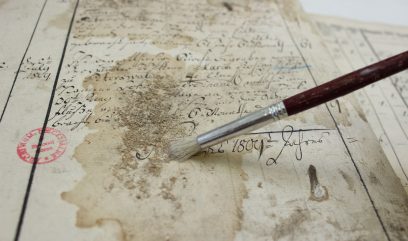 Zbliżenie na stary, poplamiony dokument z czarnymi pionowymi liniami i odręcznym pismem kursywą, czerwonym okrągłym stemplem, zabrudzeniami i fragmentami osypującego się papieru oraz pędzlem z drewnianym trzonkiem, metalowym skuwką i jasnym włosiem opartym o kartę.