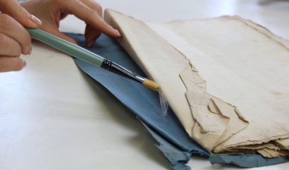 Dwie dłonie po lewej trzymają pędzelek z zielonym trzonkiem nad złożonymi, pożółkłymi i postrzępionymi kartkami leżącymi na niebieskiej podkładce na jasnym stole