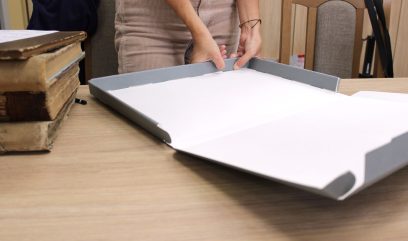 Dłonie osoby składującej szary kartonowy pojemnik z białym wkładem na drewnianym stole; obok stos starych książek, fragment beżowych sztruksowych spodni, krzesła i widoczny pasek torby w tle.