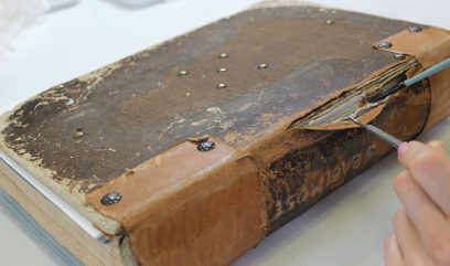 Duża, stara książka leżąca poziomo na jasnym stole z przetartą, popękaną skórzaną okładką udekorowaną metalowymi nitami i odsłoniętym, rozwarstwionym grzbietem, przy którym widać dłonie trzymające pęsetę i cienki pędzelek wsuwane między kartki, na tle rozmytych przedmiotów na stole.