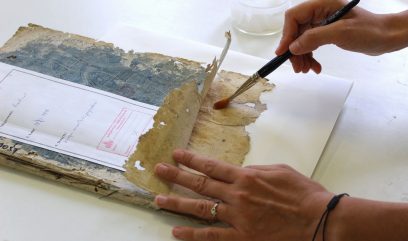 Dwie dłonie trzymają i naprawiają kruchy, pożółkły fragment papieru nad zniszczoną książką; prawa ręka trzyma pędzel nakładający wilgotną substancję, u góry stoi przezroczysty słoiczek, a na kartce widoczna biała etykieta z czerwonym stemplem.