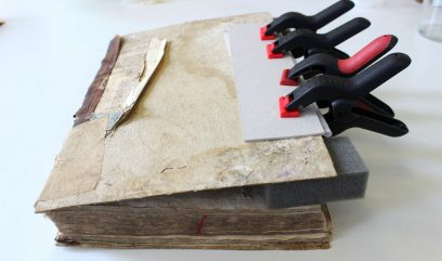 Stary, gruby tom z pożółkłą, podartą okładką i postrzępionymi kartkami leży na białej powierzchni, a przy jego górnej krawędzi prostokątną szarą płytkę przyciskają kilkoma czarnymi zaciskami z czerwonymi końcówkami opartymi na kawałku pianki.