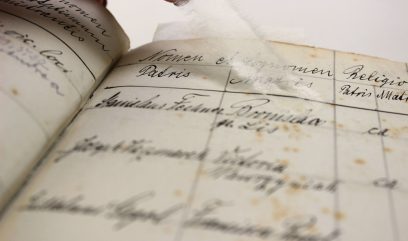 Zbliżenie na pożółkłe kartki otwartej, podniszczonej księgi z poziomymi i pionowymi liniami oraz odręcznym pismem atramentowym, z widocznymi brązowymi plamkami i kawałkiem białego włóknistego materiału umieszczonym między stronami.