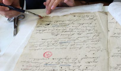 Dwie dłonie trzymają pędzel i nanoszą środek na postrzępioną krawędź pożółkłego rękopisu z odręcznym pismem i czerwonym stemplem, obok leżą metalowe nożyczki i arkusz białej bibuły.