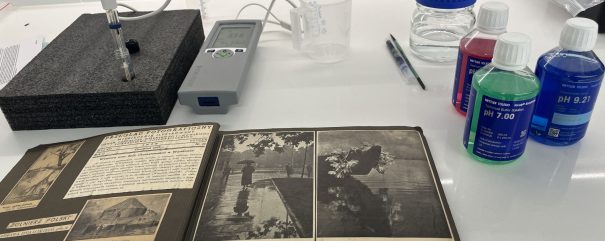 Na białym laboratoryjnym blacie otwarty stary album fotograficzny z czarno-białymi zdjęciami na pierwszym planie; obok leży przenośny miernik z wyświetlaczem i sondą osadzoną w czarnej piance, przezroczyste miarki i kilka długopisów, a po prawej plastikowe butelki roztworów buforowych o kolorowych etykietach (widoczne pH 7.00 na zielonej i pH 9.21 na niebieskiej) oraz przezroczysta butelka z niebieskim zakrętkiem.
