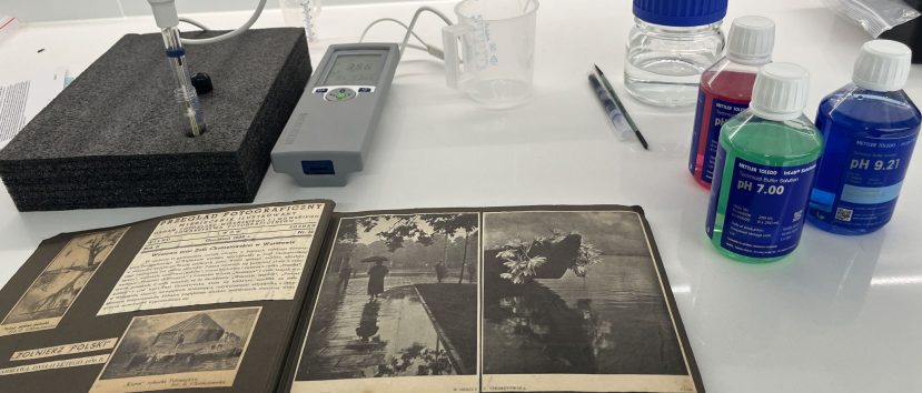 Na białym laboratoryjnym blacie otwarty stary album fotograficzny z czarno-białymi zdjęciami na pierwszym planie; obok leży przenośny miernik z wyświetlaczem i sondą osadzoną w czarnej piance, przezroczyste miarki i kilka długopisów, a po prawej plastikowe butelki roztworów buforowych o kolorowych etykietach (widoczne pH 7.00 na zielonej i pH 9.21 na niebieskiej) oraz przezroczysta butelka z niebieskim zakrętkiem.