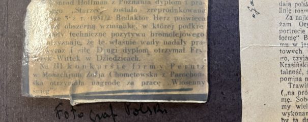 Zbliżenie ciemnobrązowego kartonu z dwoma pożółkłymi wycinkami prasowymi — po lewej mały laminowany prostokąt z częściowo przygasłym drukiem i niebieskim podkreśleniem, po prawej wąski pionowy pasek z drukowanym tekstem, na kartonie widoczny czarny odręczny zapis.