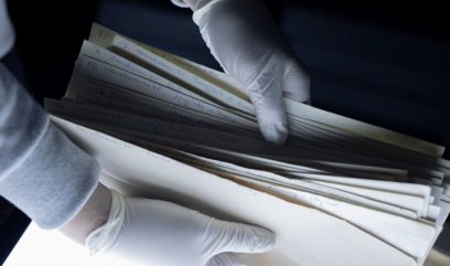 Dwie dłonie w białych lateksowych rękawiczkach trzymają i przeglądają stos papierowych kopert i kartek z odręcznymi niebieskimi zapiskami i numeracją na ciemnym tle