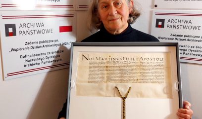 Osoba z siwymi włosami stoi przed ścianą z tablicami „Archiwa Państwowe” i trzyma oprawiony, pionowo ustawiony historyczny dokument z widocznym nagłówkiem w języku łacińskim oraz wiszącą czerwoną lakową pieczęcią na plecionym sznurze.