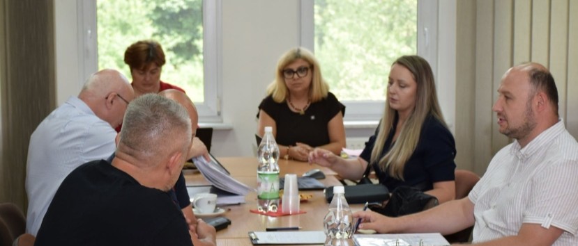 Sześć osób siedzi przy prostokątnym stole w jasnym pomieszczeniu — wokół leżą dokumenty, teczki i notatniki, są kubki oraz dwie przezroczyste butelki z wodą, w tle widać dwa okna z zielenią za nimi i pionowe żaluzje.