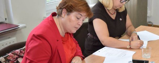 Dwie kobiety siedzą przy drewnianym stole przy oknie, pochylone nad dokumentami i podpisujące je — po lewej kobieta w czerwonej marynarce i krótkich brązowych włosach, po prawej blondynka w ciemnej sukience i okularach; na stole rozłożone kartki, notesy, długopisy, szklanki i butelki wody, a na krześle widoczna torba w kwiatowy wzór.