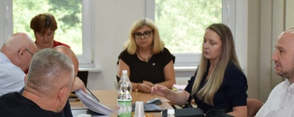 Sześć osób siedzi przy prostokątnym stole w sali spotkań — w centralnej części zdjęcia kobieta o blond włosach i okularach, po jej prawej długowłosa kobieta, pozostali mężczyźni po bokach — na stole dokumenty, segregatory, kubki i plastikowe butelki z wodą, a w tle duże okna z widokiem na zieleń.