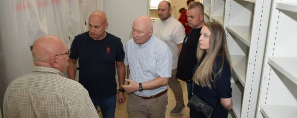Siedmioro dorosłych stoi w wąskim pomieszczeniu z pustymi metalowymi półkami po prawej i zasłoniętymi folią oknami po lewej; na pierwszym planie mężczyzna w jasnej koszuli z krótkim rękawem gestykuluje w stronę innego mężczyzny w koszuli w kratę, obok stoi kobieta w ciemnej sukience z długimi jasnymi włosami i małą czarną torebką.
