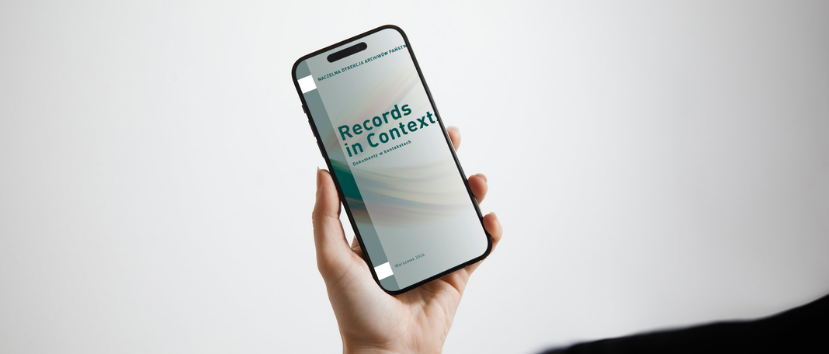 Dłoń trzyma pionowo czarny smartfon, na ekranie którego widoczna jest okładka dokumentu o zielono‑turkusowym, falistym motywie i tytułem Records in Context, na jednolitym jasnym tle z drobnym fragmentem czarnego ubrania w prawym dolnym rogu.
