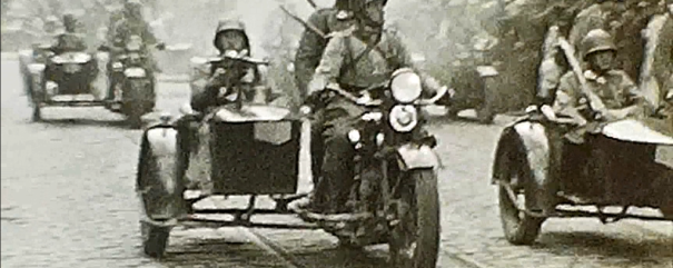 Czarno‑białe zdjęcie grupy żołnierzy w hełmach jadących motocyklami z bocznymi wózkami i widocznymi karabinami po brukowanej ulicy z torami tramwajowymi, w tle inne pojazdy i zgromadzeni ludzie.