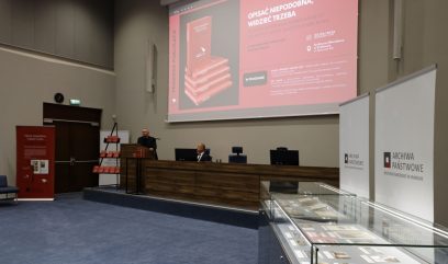 Duża sala konferencyjna z niebieskim dywanem, drewnianą mównicą i stołem prelegentów, w której dwóch mężczyzn przy mównicy i za stołem prowadzi prezentację na dużym ekranie z czerwonym slajdem pokazującym stos książek i tekst, z przodu szklana witryna z dokumentami oraz po bokach stojące roll-upy i plakaty z napisem Archiwa Państwowe.