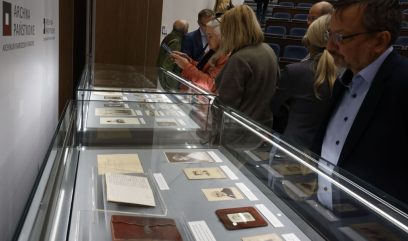 Grupa osób przygląda się przeszklonym gablotom z ułożonymi starymi dokumentami, zdjęciami i notesami w sali wystawowej; po lewej na ścianie widoczny napis 