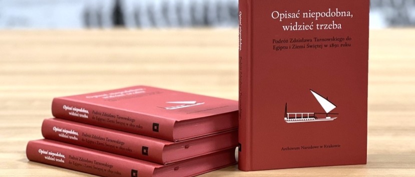 Na drewnianym stole trzy czerwone książki ułożone poziomo obok jednej stojącej okładką do przodu; na okładkach biały tytuł 